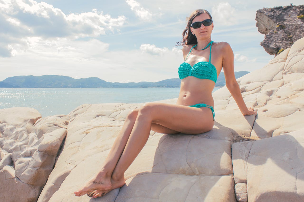 A woman wearing sunglasses and a bikini relaxes on a rocky seashore with mountains in the distance.