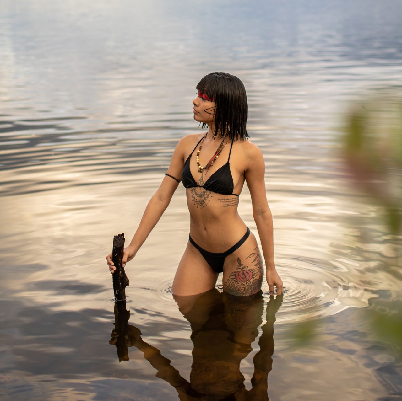 A woman with tattoos standing in a lake wearing a bikini, holding a stick.